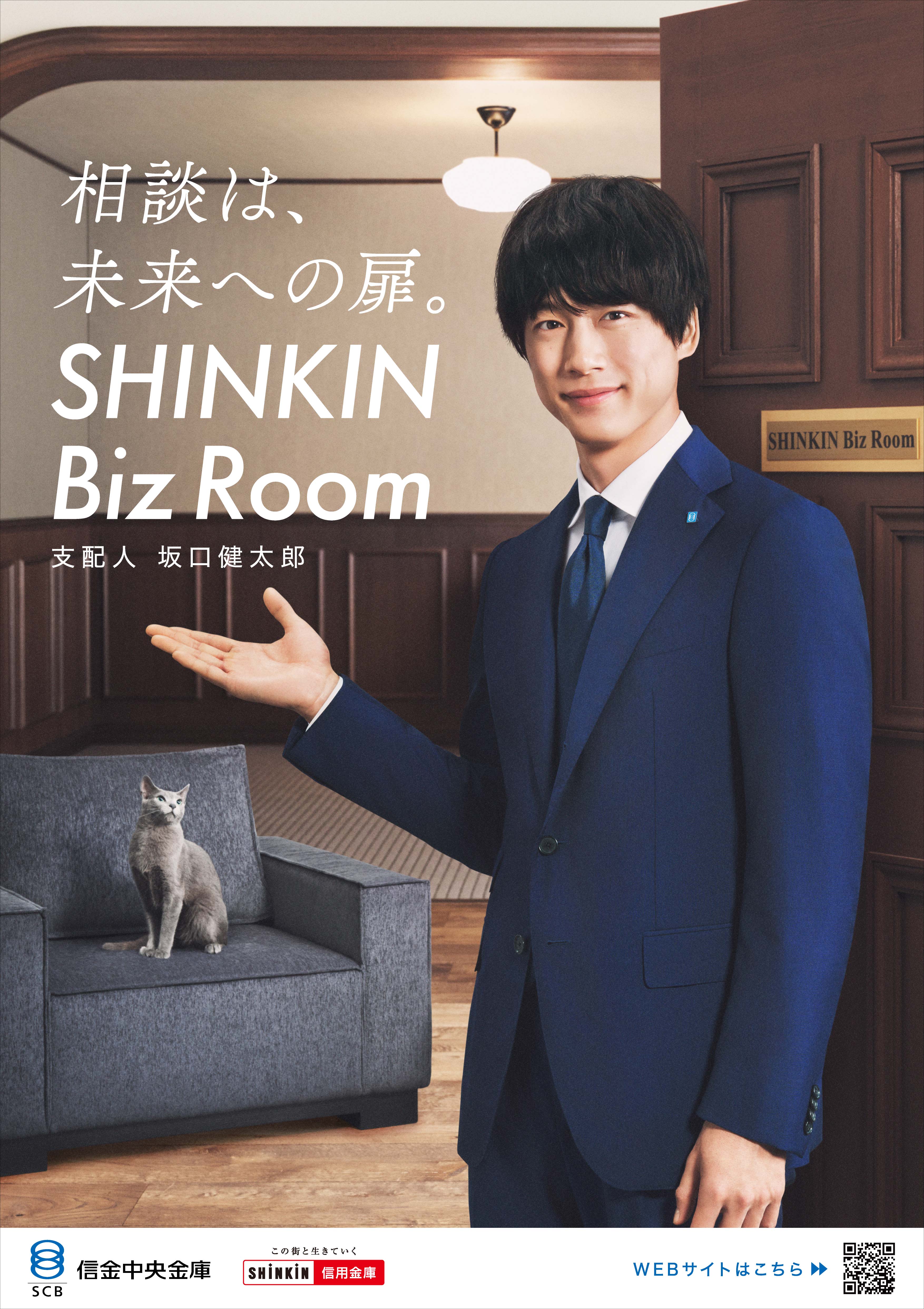 相談は、未来への扉。SHINKIN Biz Room 支配人 坂口健太郎 スーツ姿の坂口健太郎が右手を差し出し、背後のソファに座る灰色の猫を指し示している。背景は木の壁と扉がある落ち着いた室内。