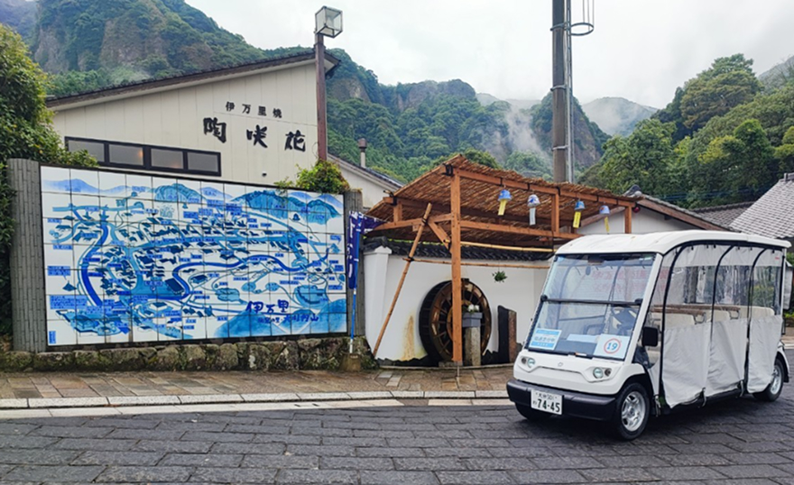 屋根付きの小型電動カート（透明サイドカーテン付き）が道路脇に停車。背景に観光案内の看板や建物が見え、地域の移動手段として活用されるイメージ。