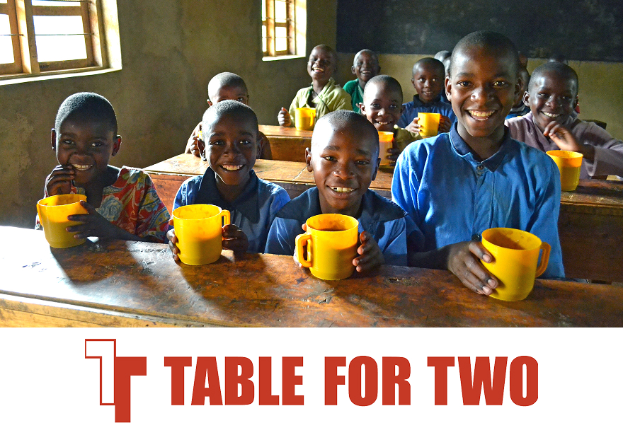 教室の机に並ぶ数人の子どもが黄色いマグを持ってこちらに笑顔を向ける。途上国の子どもの食支援を示す「TABLE FOR TWO」ロゴ付き。